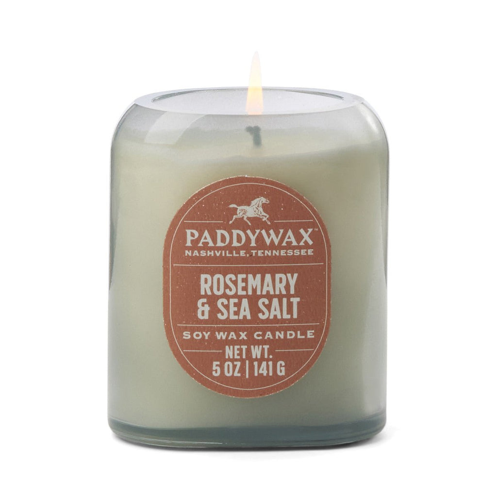 The Paddywax - Vista Rosemary & Sea Salt candle in a denim blue glass jar, weighing 5 oz (141 g), is labeled Paddywax, Nashville, Tennessee. Its warm glow reflects American West fragrances spirit with captivating scents.