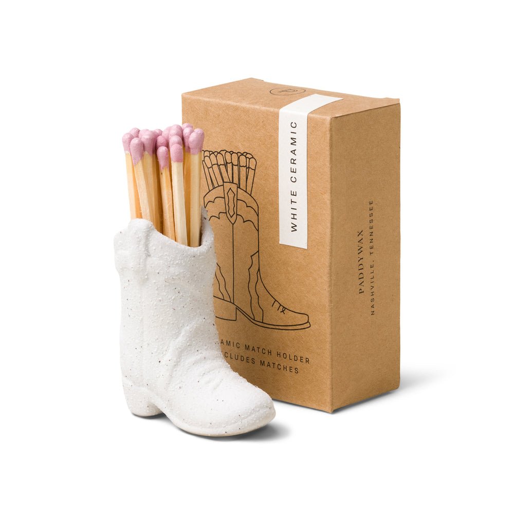 A vintage-style ceramic holder shaped like a cowboy boot, made by Paddywax, contains pink-tipped matches. It stands next to a brown box labeled Paddywax - White Cowboy Boot Match Holder, featuring an illustration of the boot and indicating it includes matches.