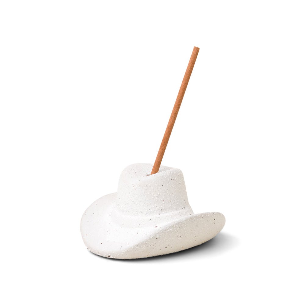 The Paddywax white ceramic cowboy hat incense holder features a textured design with an incense stick inserted at the top, slightly angled to the right, evoking Nashvilles charm. It casts a subtle shadow on a plain white background.