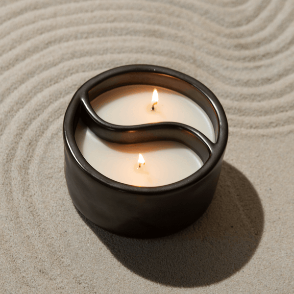 The Paddywax Yin-Yang Palo Santo + Cade 11oz Candle, with its black ceramic design and dual flame feature, rests calmly on raked sand. Its wax is divided by a wave-like pattern, creating two sections that emit unique fragrances, enhancing the zen atmosphere and inviting inner peace.