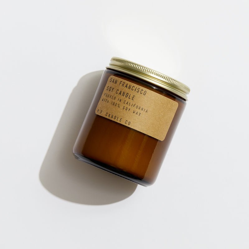 The P.F. Candle Co. - 7.2 oz. San Francisco Candle features an earthy scent in a brown glass jar with a gold metal lid, casting a soft shadow on a white backdrop.