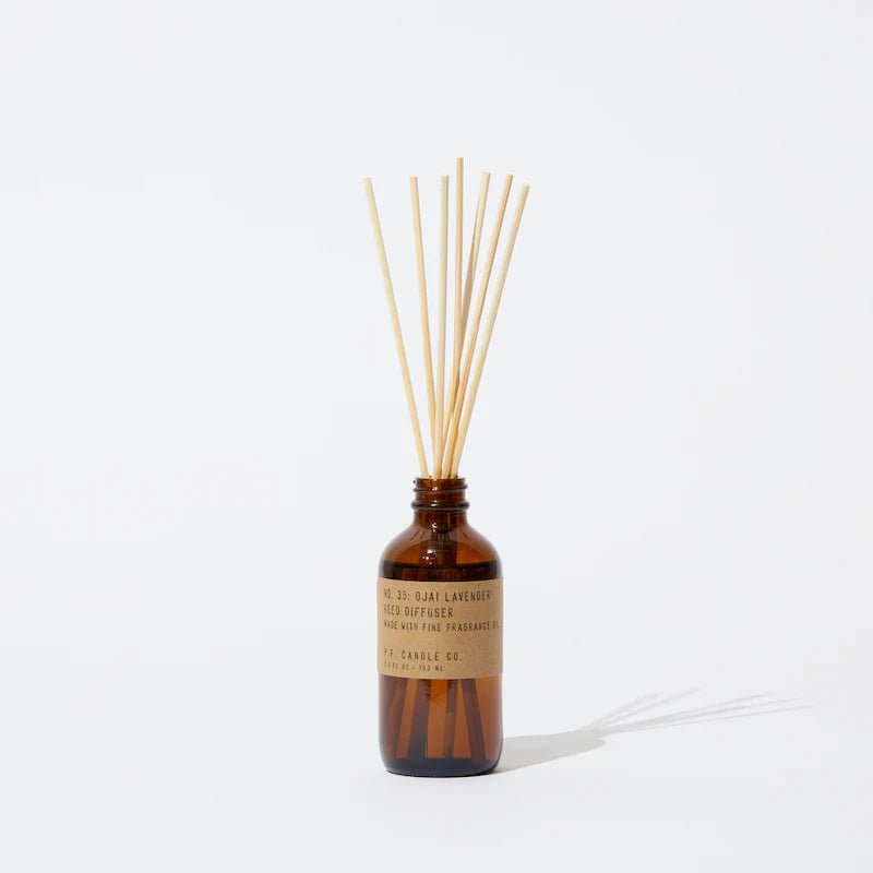 A brown glass bottle labeled P.F. Candle Co. - Diffuser - Ojai Lavender holds slender reed sticks, suggesting a reed diffuser with a fresh, earthy fragrance. Set against a plain white background, the bottles subtle shadow evokes the calming essence of Ojai Lavender.