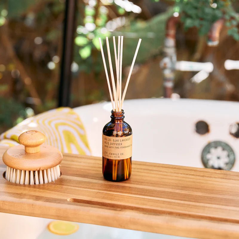 A P.F. Candle Co. Ojai Lavender diffuser in a small glass bottle enhances the serene bathroom vibe, resting on a wooden tray beside a round-bristle body brush, with green plants and a bathtub completing the setting.