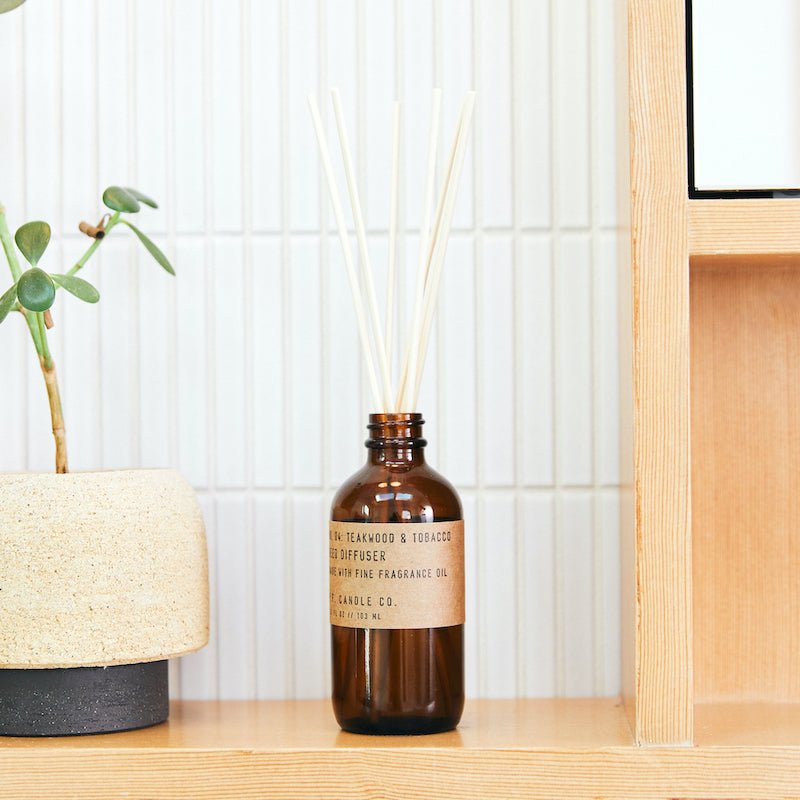 A brown glass bottle labeled P.F. Candle Co. - Diffuser - Teakwood and Tobacco emits a subtle teak scent on a light wooden shelf, with white reeds fanning gracefully. A potted plant in a beige pot complements the look against a white textured wall.