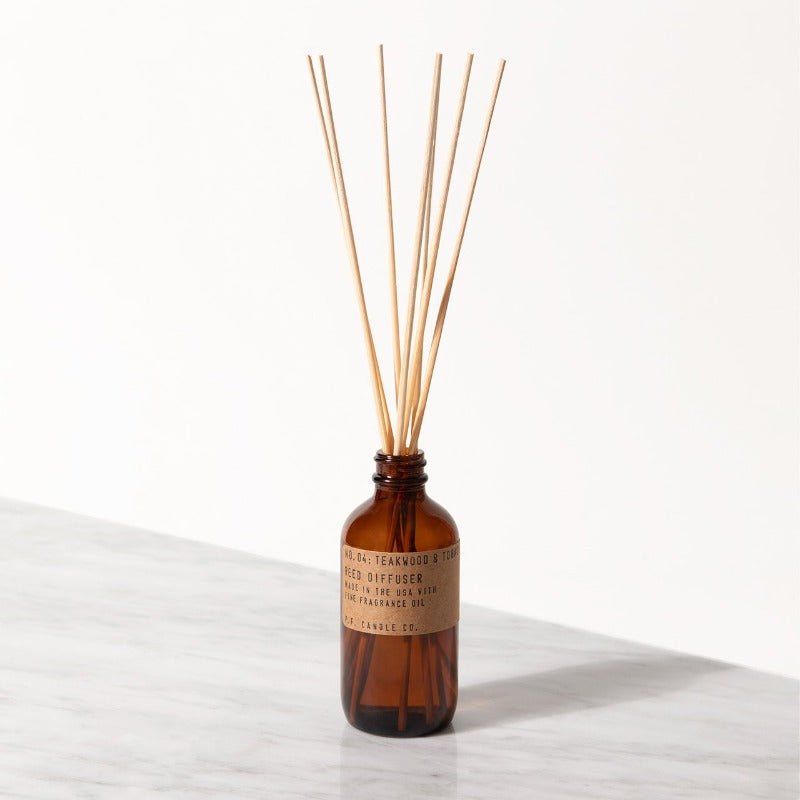 The P.F. Candle Co. - Diffuser, a teakwood and tobacco-scented brown glass bottle with a label, releases fragrance through thin wooden reeds on a white marble surface against a neutral backdrop.