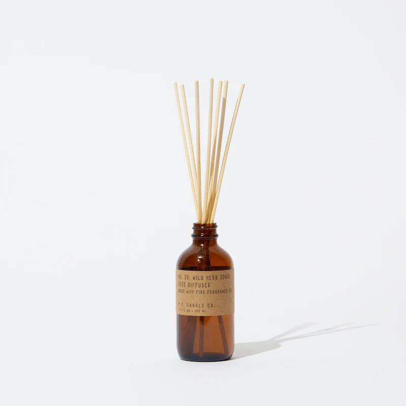 A brown glass P.F. Candle Co - Diffuser labeled Wild Herb & Tonic stands upright, with thin reed sticks emerging from the top, filling the air with an aromatic and woody scent while casting a soft shadow on a light background.