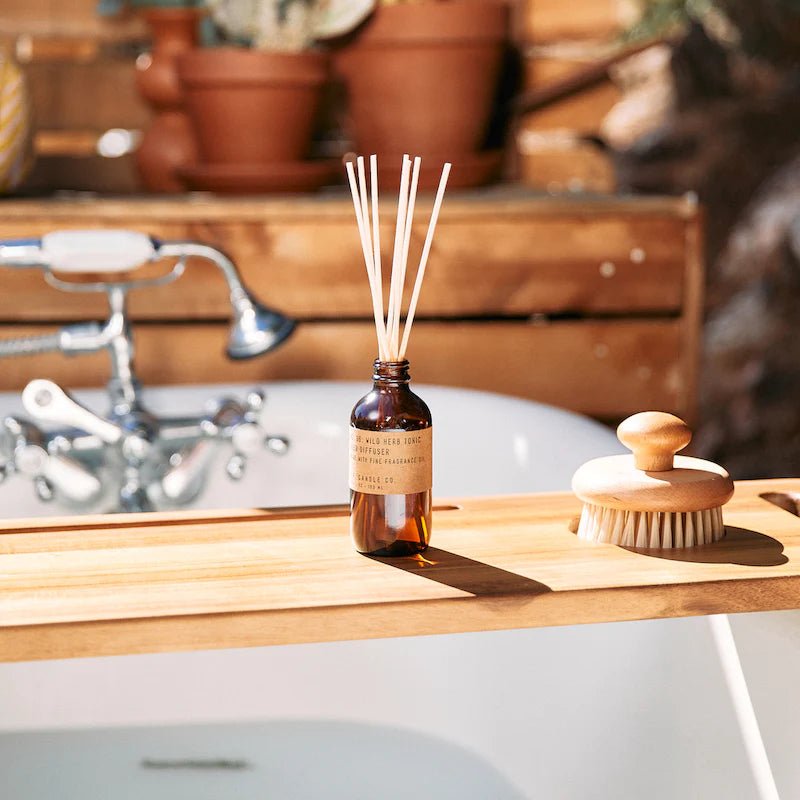 A wooden bath tray spans the tub, holding a P.F. Candle Co Diffuser in Wild Herb & Tonic and a body brush. Sunlight highlights the rustic bathroom with potted plants and a vintage-style faucet backdrop.