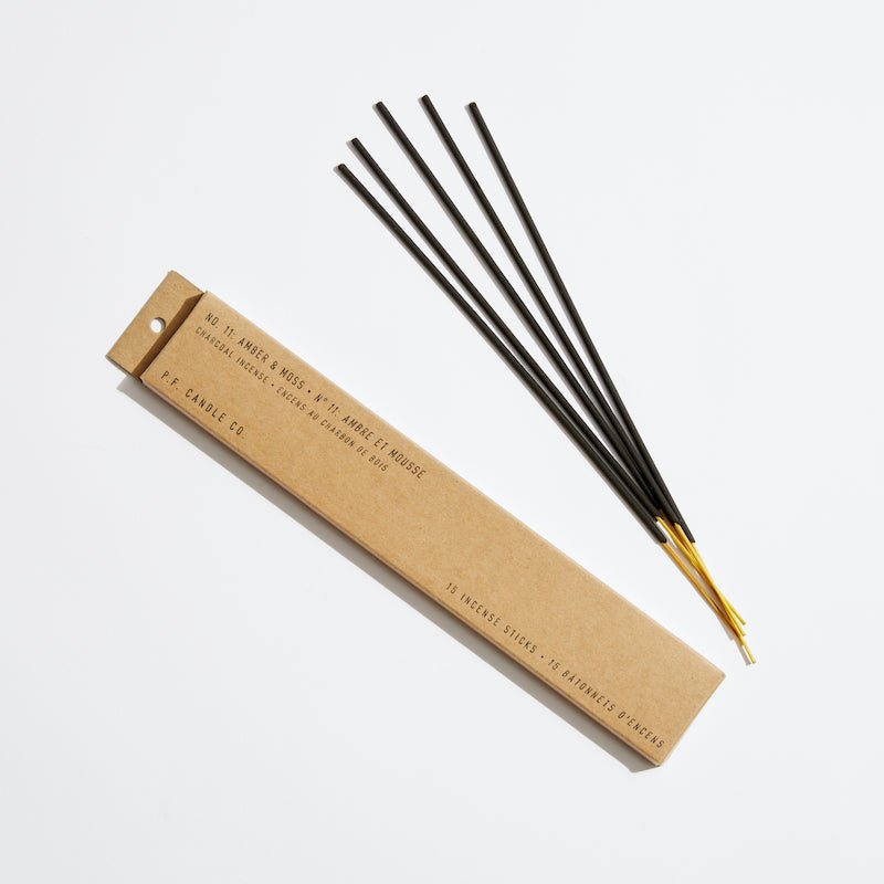 A brown P.F. Candle Co. box is placed next to four black incense sticks with golden tips on a white backdrop. The box is labeled No. 11 Amber & Moss and contains 15 Hand-dipped Incense Sticks, offering a rich fragrance experience.