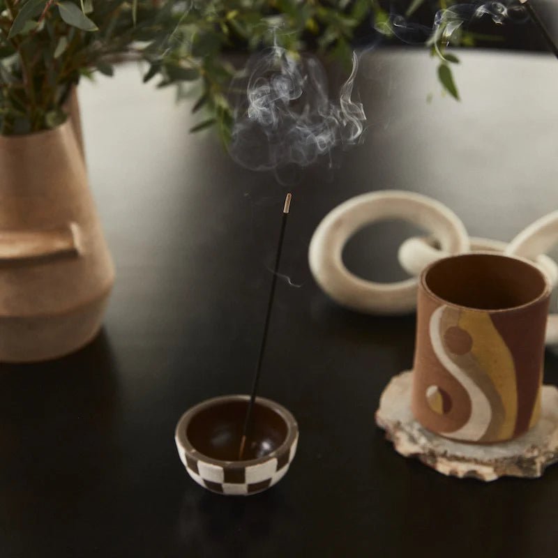 A P.F. Candle Co. Piñon incense stick releases a woody aroma from a checkered holder on a dark surface. Nearby, a yin-yang mug and decorative pottery complement the greenery, enhancing the serene setting.