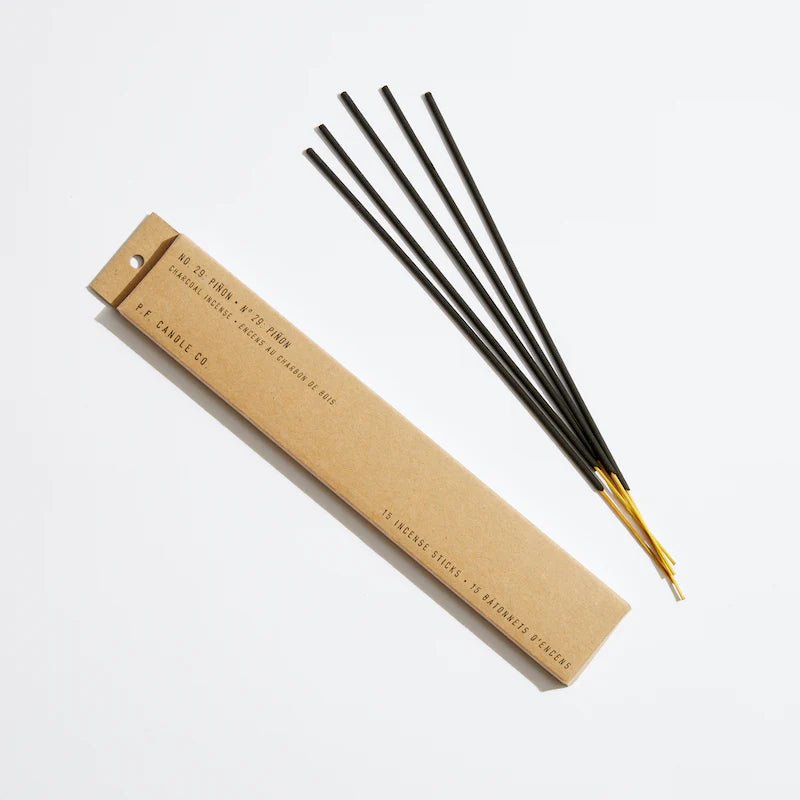 A white box labeled No. 29: Piñon from P.F. Candle Co. holds 15 incense sticks, emitting a woody aroma with each black stick burning for 45 minutes. Five sticks are placed beside the packaging, accenting the minimalist design against a plain white background.