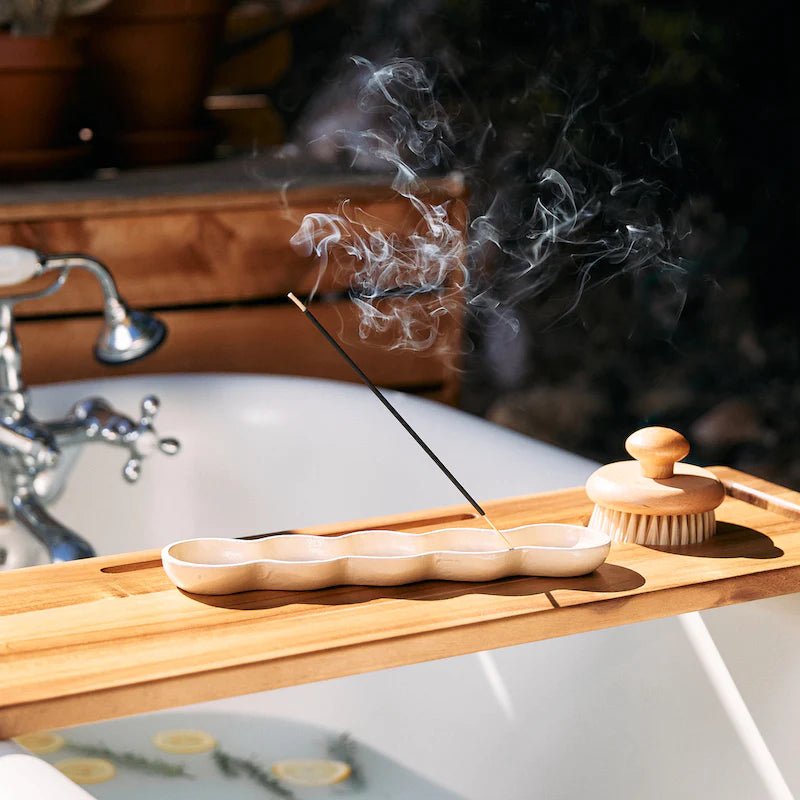 A wooden bath caddy sits across a white bathtub for an aromatic ambiance. The P.F. Candle Co.s Wild Herb Tonic incense stick emits smoke from its holder, paired with a wooden bath brush. The tub is filled with water and lemon slices for total relaxation.