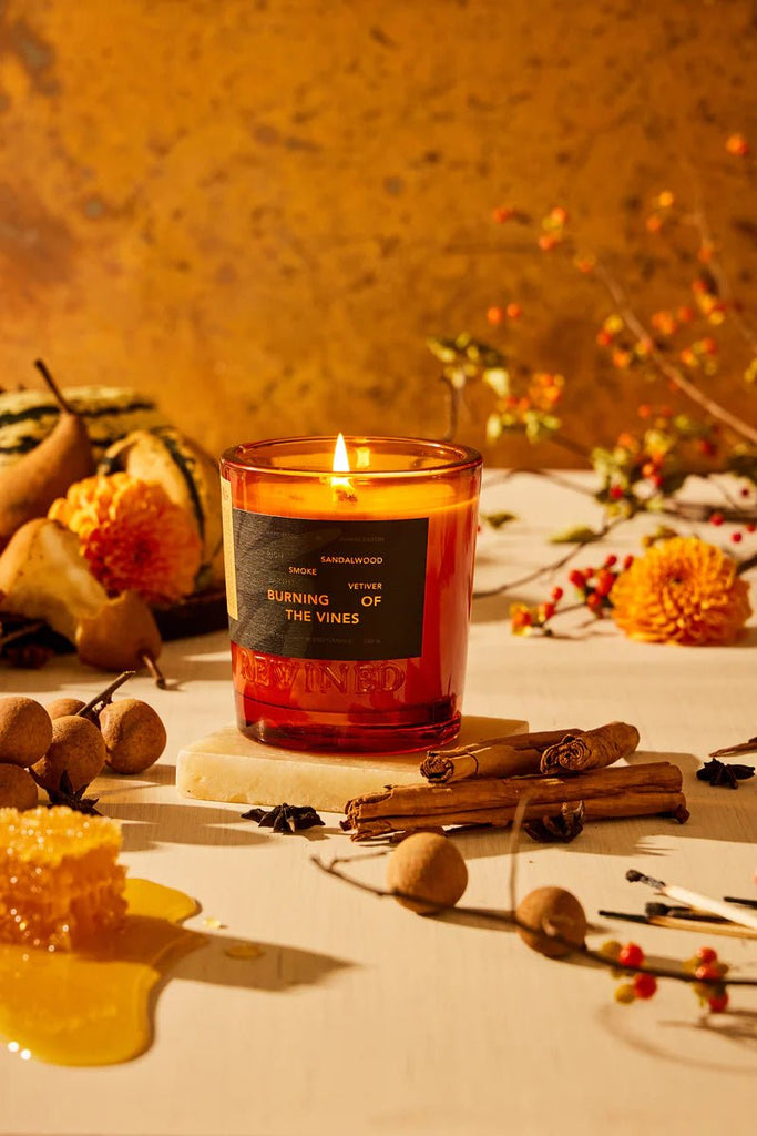 A lit Rewined Burning Of The Vines 6oz candle is surrounded by cinnamon sticks, yellow flowers, fruits, and honeycomb on wood. The warm-textured wall with autumnal decor enhances the inviting fragrance ambiance.