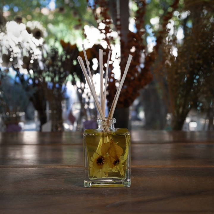 The Rosy Rings Botanical Diffuser in Honey Tobacco features a glass bottle filled with yellow liquid and flower petals, topped with white reed sticks. Positioned on a wooden table against a blurred backdrop of dried flowers and a sunlit window, it subtly hints at its soothing fragrance notes.