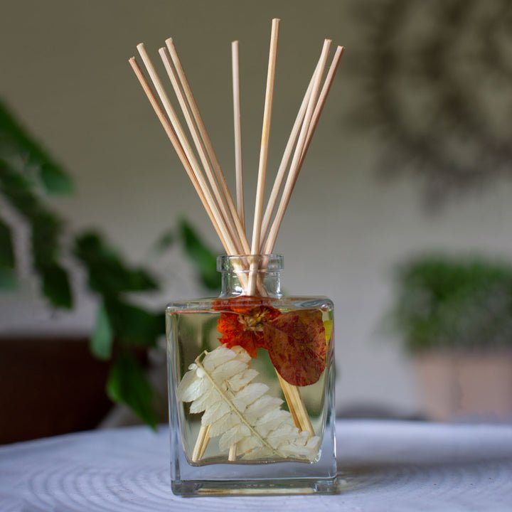 A square glass bottle with light wooden reed sticks contains the Rosy Rings Botanical Diffuser in Honey Tobacco. Dried leaves and orange petals inside emit a subtle fragrance, set against a blurred background with a green plant, enhancing the natural ambiance.