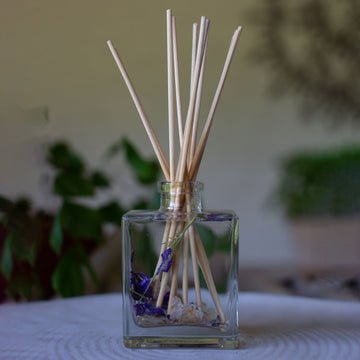 The Rosy Rings Botanical Diffuser, Roman Lavender 4oz, features a square glass bottle filled with light-colored scented sticks and showcases dried purple flowers, with lavender botanicals hinting at soothing fragrance notes against a softly blurred green foliage background on a white surface.