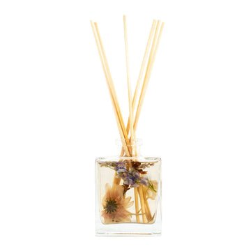 The Rosy Rings - Botanical Diffuser in Roman Lavender features a clear glass bottle filled with liquid and dried lavender. Wooden reeds extend from the top, while purple and peach flowers provide a natural, decorative look to enhance your spaces subtle fragrance notes.