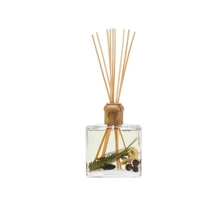 The Rosy Rings - Forest 13oz Botanical Diffuser features a clear bottle with a wooden cap that holds liquid and thin reeds. Delicate botanicals like pine sprigs and cones enhance the natural aesthetic, offering a calming, invigorating cedarwood atmosphere.