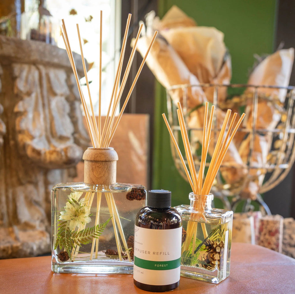 Two Rosy Rings Forest 13oz Botanical Diffusers with natural elements and wooden tops sit on a surface. A small bottle labeled Diffuser Refill Forest releases cedarwood and botanicals, set against a backdrop of an abstract interior with decorative items.