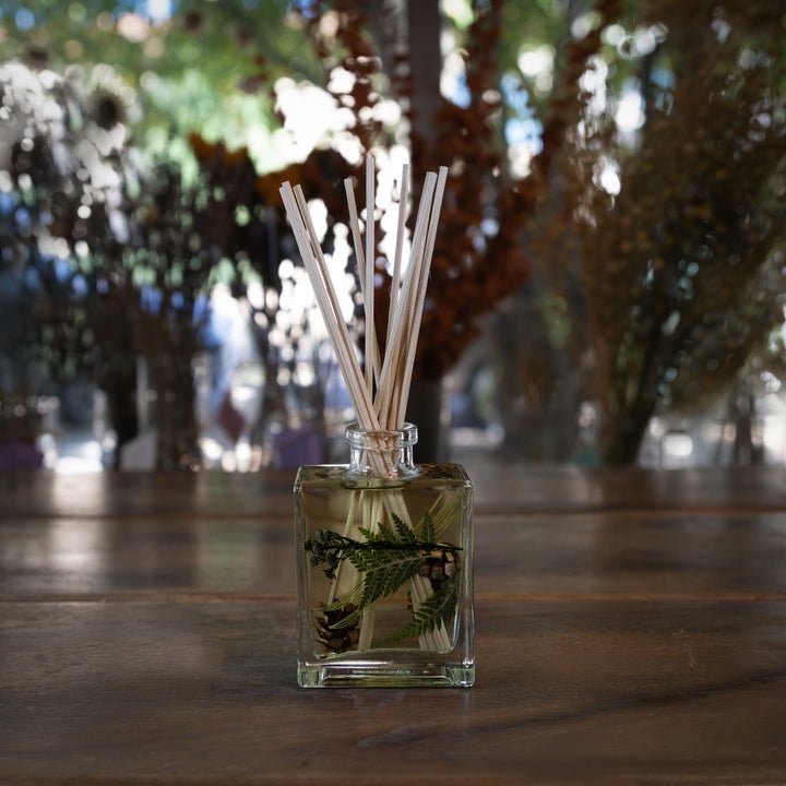 A Rosy Rings - Forest 4oz Botanical Diffuser sits on a wooden table, reed sticks rising to fill the air with fragrance. In the background, blurred plants and dappled light from trees complete the serene setting.