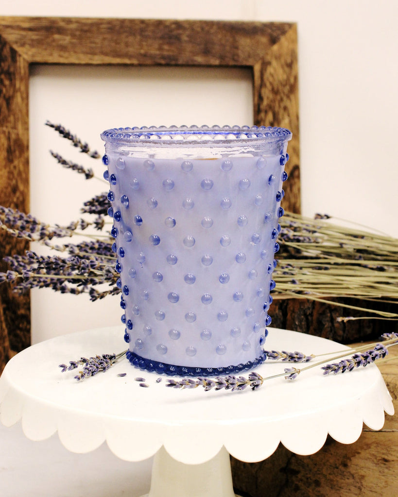 A Simpatico Lavender Hobnail Candle, made from soy wax and essential oils, is on a white pedestal stand with dried lavender stems around it. A wooden frame leans against a light wall in the background.