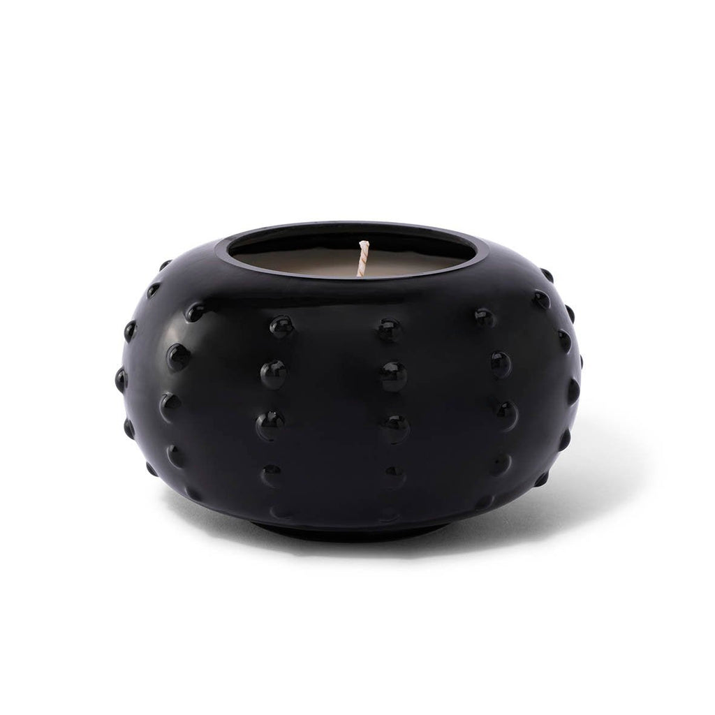 The Skeem - Black Candlescape 10 oz Candle in Tobacco Noir features a round black holder with a textured, dotted surface and a single white wick. This candle is perfect for creating a cozy, holiday ambiance against its plain white background.