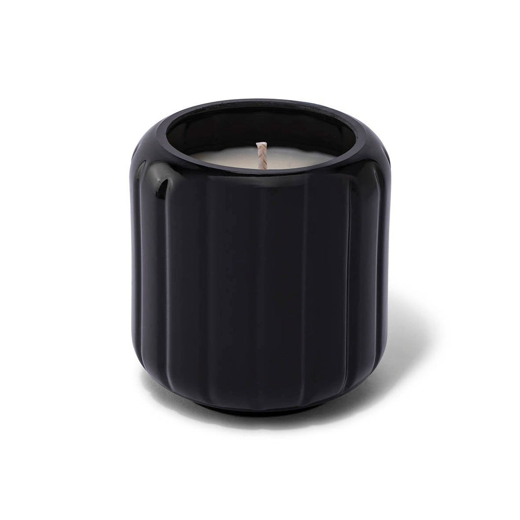 A black ribbed ceramic holder with a white candle evokes a Tobacco Noir ambiance against a plain backdrop. This Skeem Black Candlescape 8 oz Candle is perfect for infusing your space with rich, holiday scents.