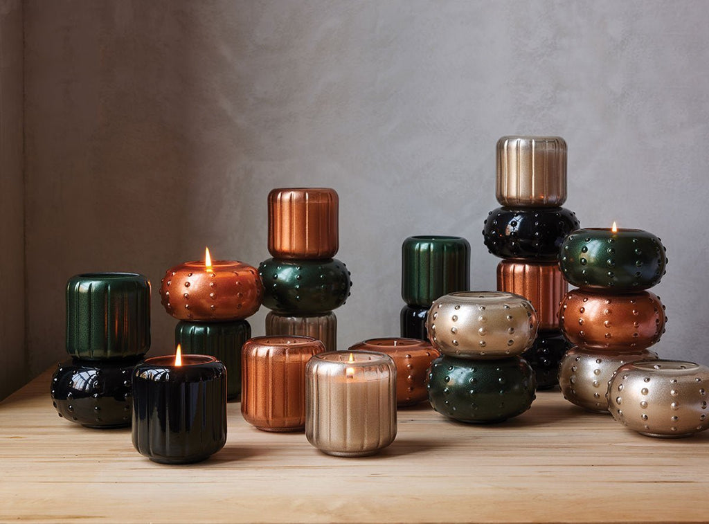 Skeems Champagne Candlescape showcases decorative Yuzu Pine-scented candles in textured holders atop wood. These candles, in metallic hues like copper, gold, black, and green, emit a warm glow or rest stacked against a neutral wall to create an elegant display.