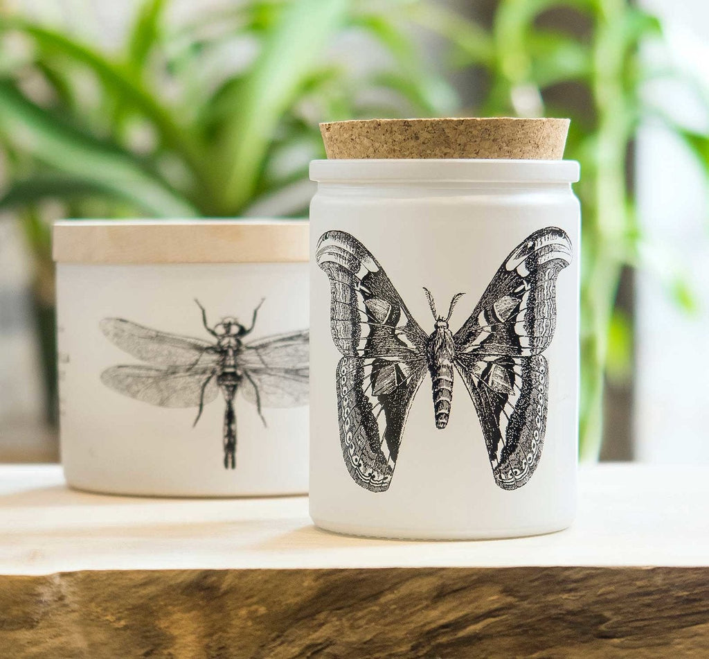 The Skeem - Citronella Sea Salt Candle, in a white jar with a cork lid and black butterfly illustration, contains soy-based citronella oil. Nearby, another Skeem white container with a dragonfly design is seen among blurred green plants.