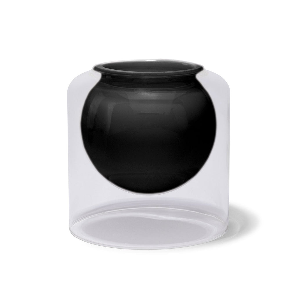 The Skeem - Fig Noir Luna 20oz Candle exudes luxury with its modern design, featuring a transparent glass layer and a black, spherical inner chamber. Its minimalist cylindrical shape stands gracefully on a flat base, adding elegance to any space.