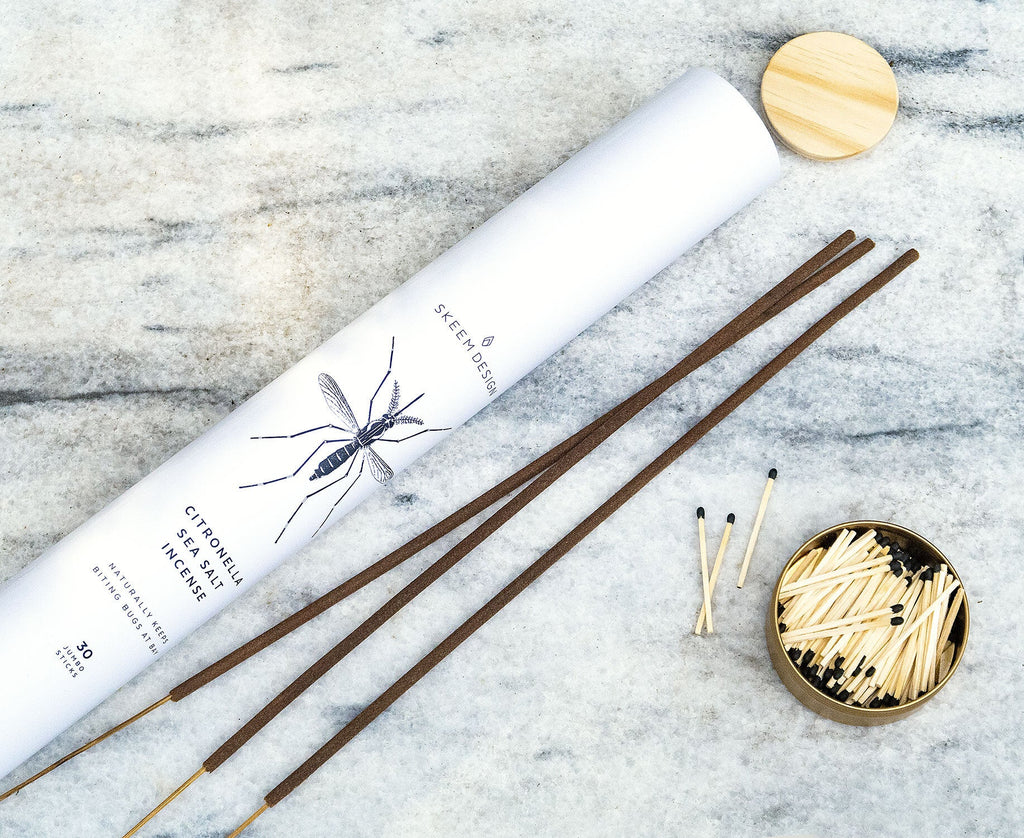 A Skeem - Jumbo Citronella Sea Salt Incense tube, featuring a mosquito graphic, rests on marble amid jumbo incense sticks and an open tin of matches—ideal as a natural insect repellent.