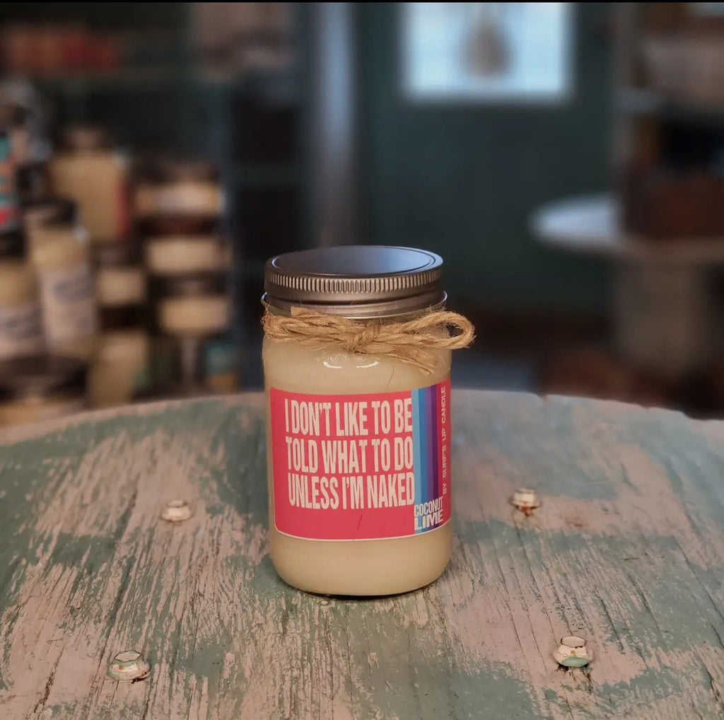 The Surf’s Up Be Told Coconut Lime Mason Jar Candle from Surfs Up Candles, featuring a humorous I dont like to be told what to do unless Im naked label, sits on a rustic green table. Made with natural soy wax and Coconut Lime scent, it charms the cozy, dimly lit room.