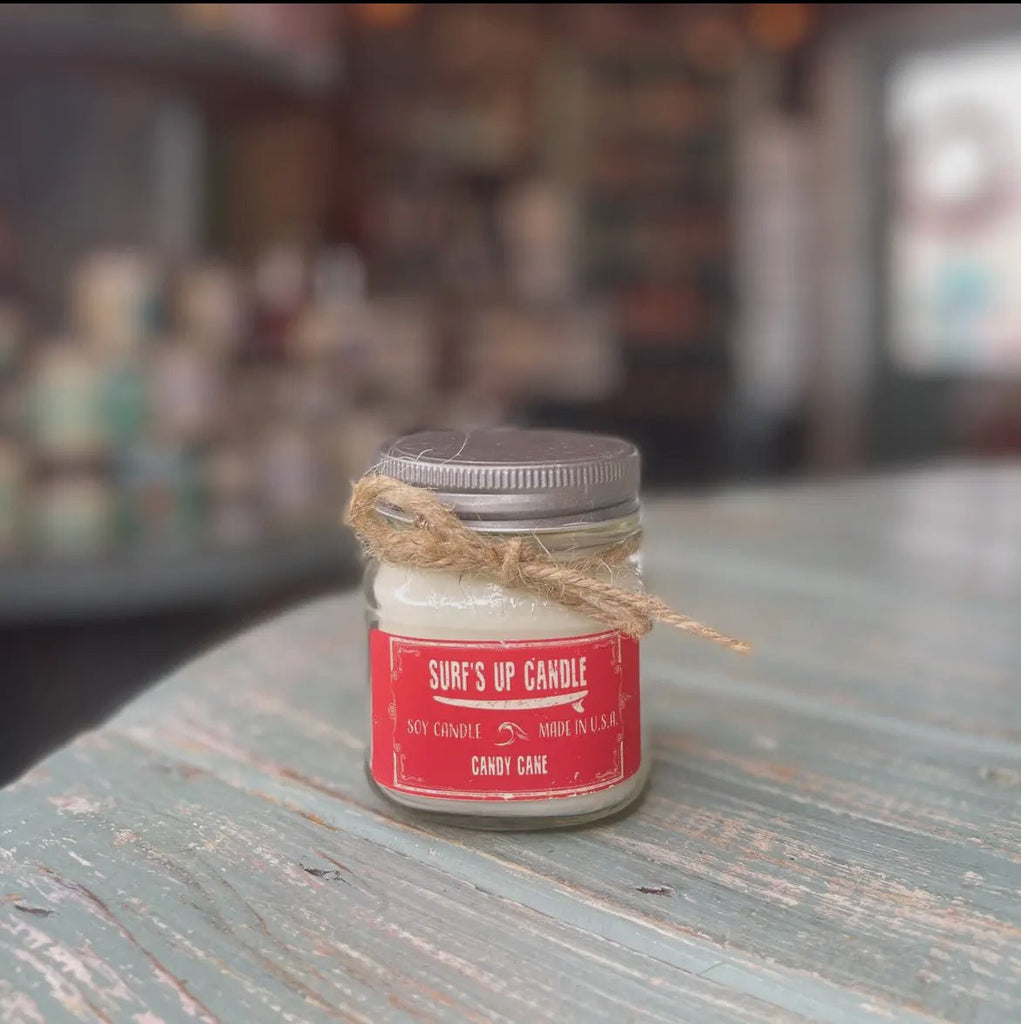 A small Surfs Up - Candy Cane Mason Jar Candle, made from soy wax with a red sticker and twine around the lid, sits on a rustic wooden table. The blurred background adds to the cozy ambiance, perfect for the peppermint and vanilla scent by Surfs Up Candles to dazzle your senses.