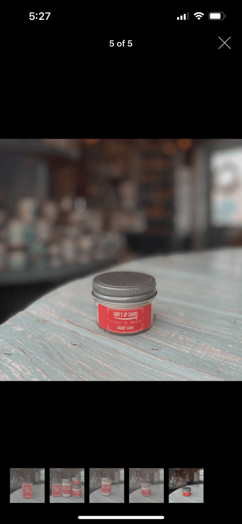 A small jar with a red label saying Surf’s Up - Candy Cane Mason Jar Candle sits on a light blue wooden surface, hinting at indulgent peppermint and vanilla scents. In the blurred background, soy wax candles and other cozy items fill the shelves, all from Surfs Up Candles.