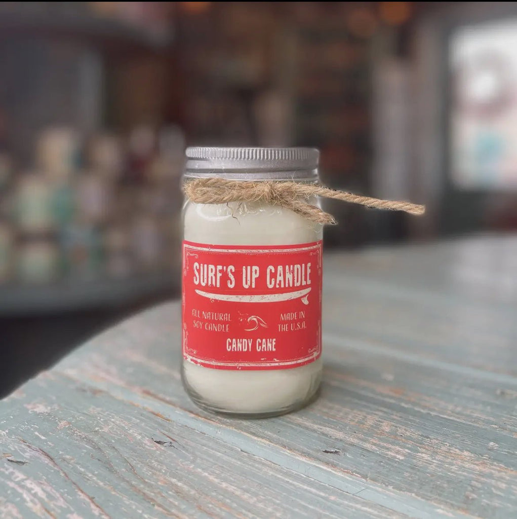 The Surf’s Up - Candy Cane Mason Jar Candle by Surfs Up Candles features a red label, peppermint and vanilla scent, and is crafted from soy wax. Tied with twine around its neck, it sits on a weathered wood surface with a softly blurred background.