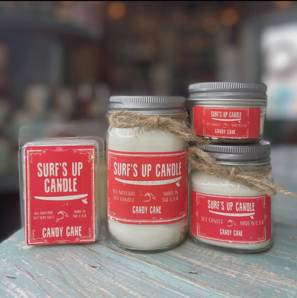 Four Surf’s Up - Candy Cane products by Surfs Up Candles are displayed, featuring a Peppermint and Vanilla scent. The set includes a soy wax melt pack and three candles in various jars, each with red labels and twine accents, elegantly arranged on a rustic wooden surface.