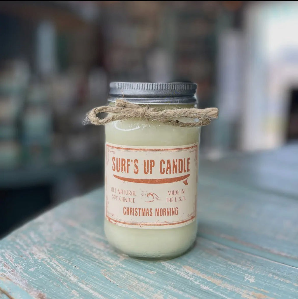 A white Surf’s Up - Christmas Morning candle by Surfs Up Candles, made of all-natural soy wax and encased in a mason jar with a metal lid and twine bow, is labeled Made in the U.S.A. and rests on weathered wood.