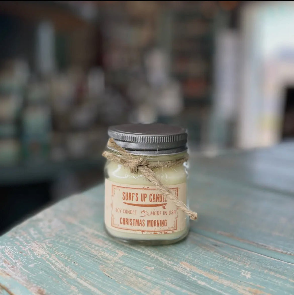 The Surf’s Up - Christmas Morning Mason Jar Candle by Surfs Up Candles, made from all-natural soy wax, sits on a rustic blue-green wooden surface. This cream-colored candle comes with a silver lid and twine around the neck, in a reusable mason jar. Its blurred background elevates its charm.