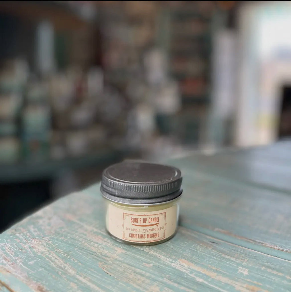 A small Surf’s Up - Christmas Morning Mason Jar Candle, made of all-natural soy wax and housed in a reusable mason jar, sits on a rustic light blue wooden table. The softly blurred background showcases shelves filled with items evoking festive warmth.