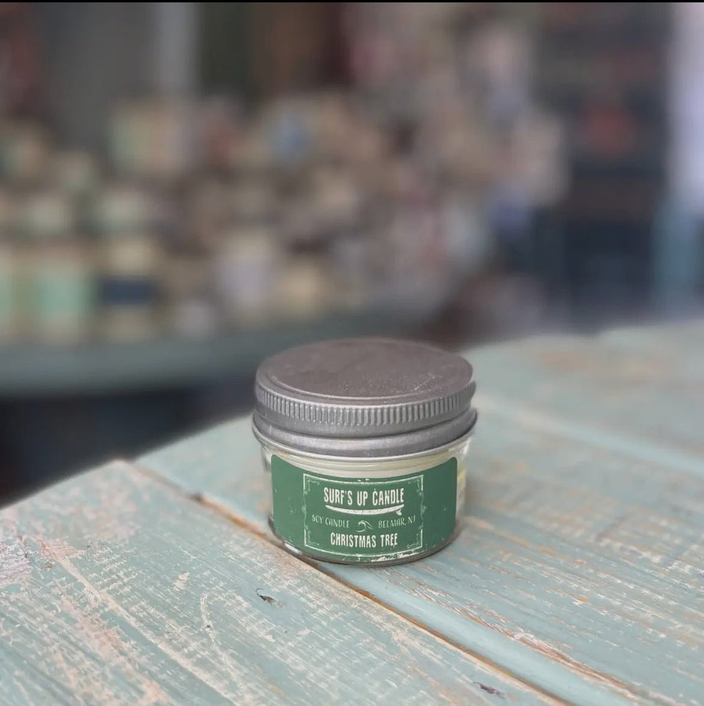 A small glass jar candle with a metal lid sits on a weathered wooden surface. The label reads Surf’s Up - Christmas Tree Mason Jar Candle, featuring natural soy wax for a cleaner burn. In the blurred background, assorted reusable Mason Jar candles from Surfs Up Candles can be seen.