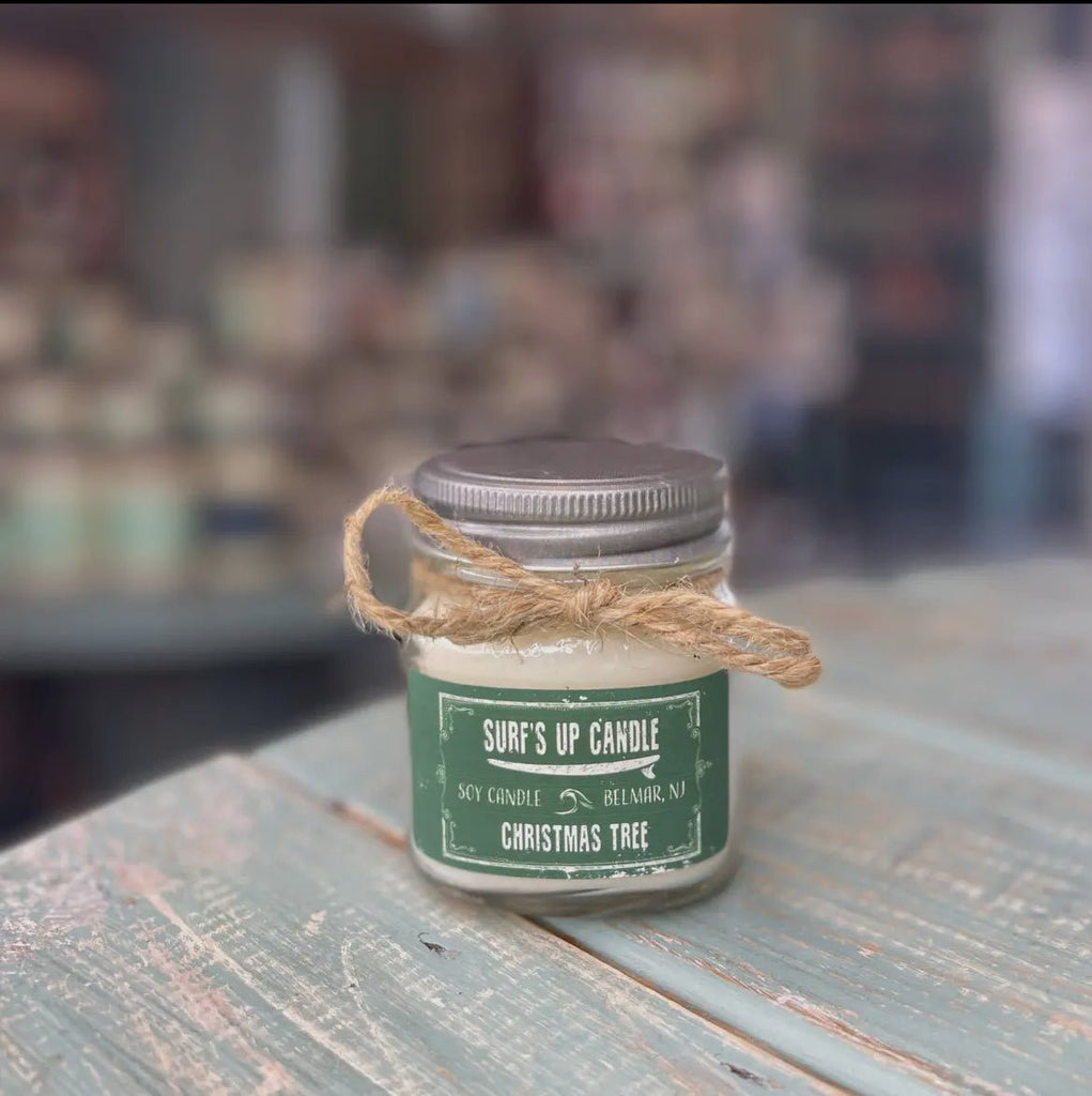 A small Mason jar candle with a metal lid sits on a rustic blue table. Made from natural soy wax, it features a green label: Surf’s Up Candle, Belmar, NJ, Christmas Tree. A twine bow adorns its neck. Background is blurred. Brand: Surfs Up Candles.