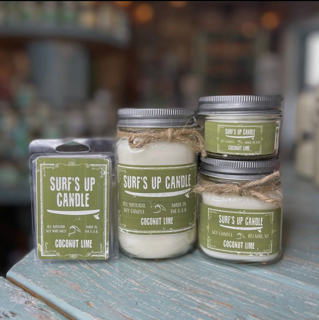 Surf’s Up - Coconut Lime Mason Jar Candle by Surfs Up Candles crafted from natural soy wax on a wooden surface, includes two reusable Mason jars with twine and a wax melt pack, all with green labels; blurred product shelves in the background.