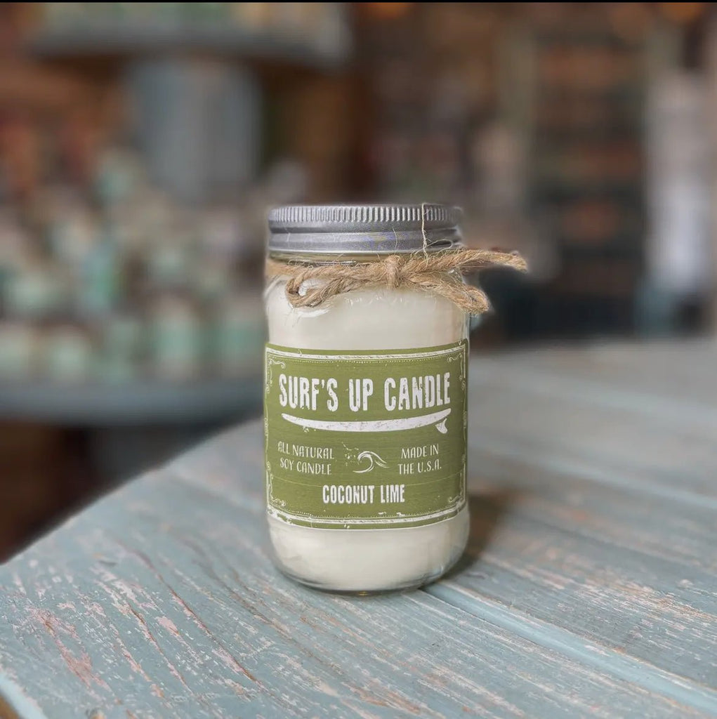 A Surf’s Up - Coconut Lime Mason Jar Candle by Surfs Up Candles sits on a rustic blue wooden surface. Made with all-natural soy wax and featuring a coconut lime scent, it has twine around the neck for added charm against its blurred background.