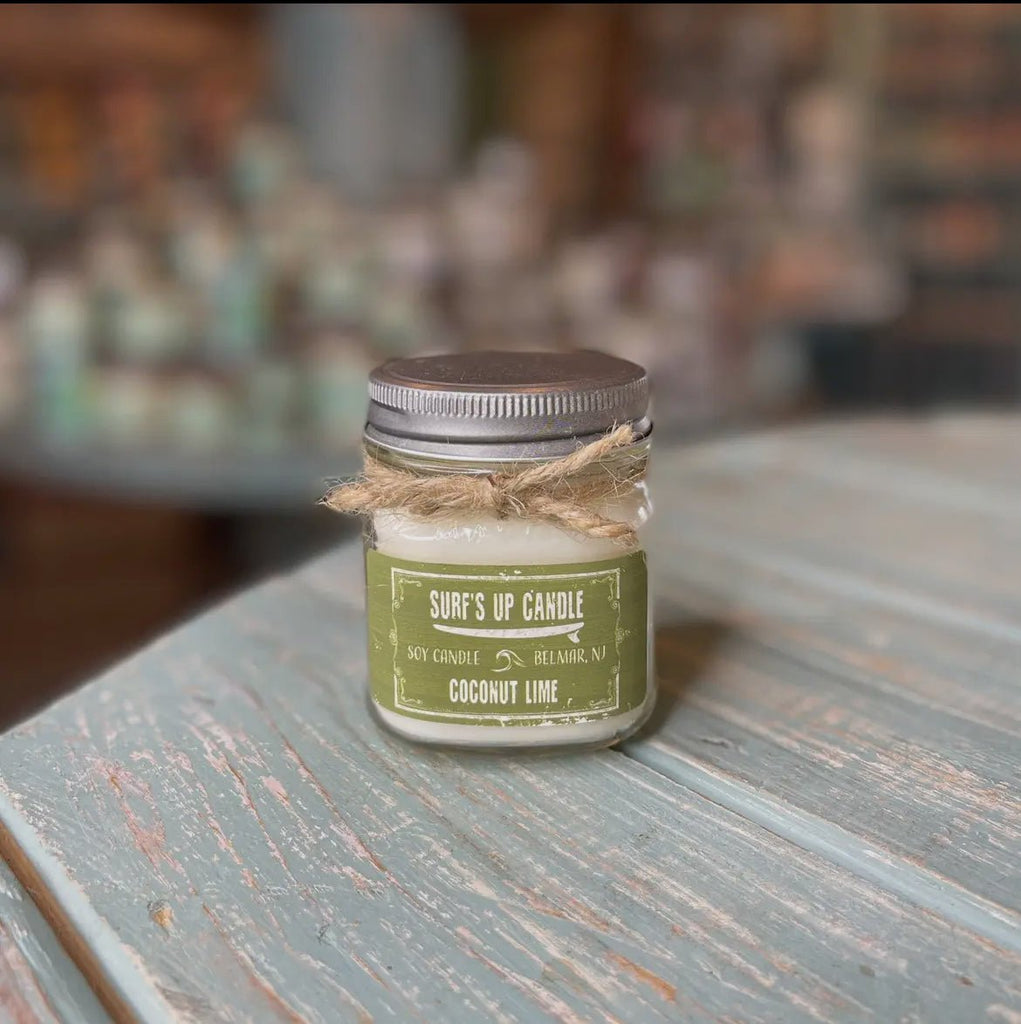 The Surf’s Up - Coconut Lime Mason Jar Candle by Surfs Up Candles is crafted from natural soy wax, adorned with a twine bow, and offers a refreshing scent. It sits on a rustic, light blue wooden table with a blurred background.