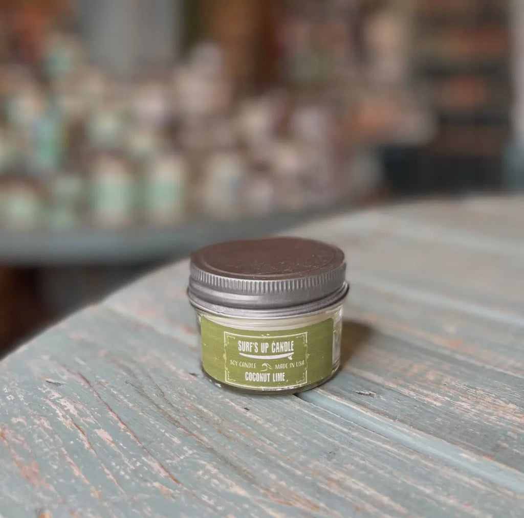 A small Surfs Up - Coconut Lime Mason Jar Candle by Surfs Up Candles with a brown lid sits on a weathered blue wooden surface. Made from natural soy wax, its label is visible against a blurred background of shelves, reflecting its reusable Mason Jar charm.