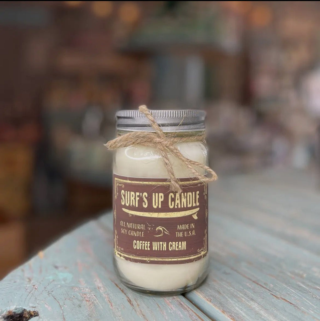 A Surfs Up - Coffee With Cream Mason Jar Candle by Surfs Up Candles sits on a weathered wooden table. The label reads All Natural Soy Candle, suggesting its coffee fragrance, while a rustic rope adorns the lid of the reusable mason jar against a softly blurred background.