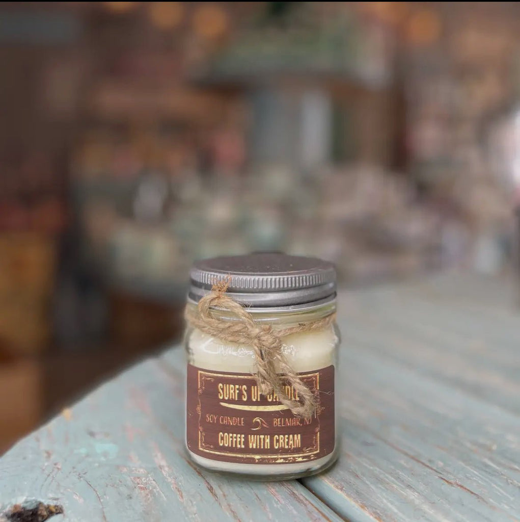 A Surfs Up - Coffee With Cream Mason Jar Candle by Surfs Up Candles sits on a rustic, weathered wooden table. Made from natural soy wax and tied with twine, it contrasts beautifully against the blurred cozy interior, emphasizing its reusable mason jar.