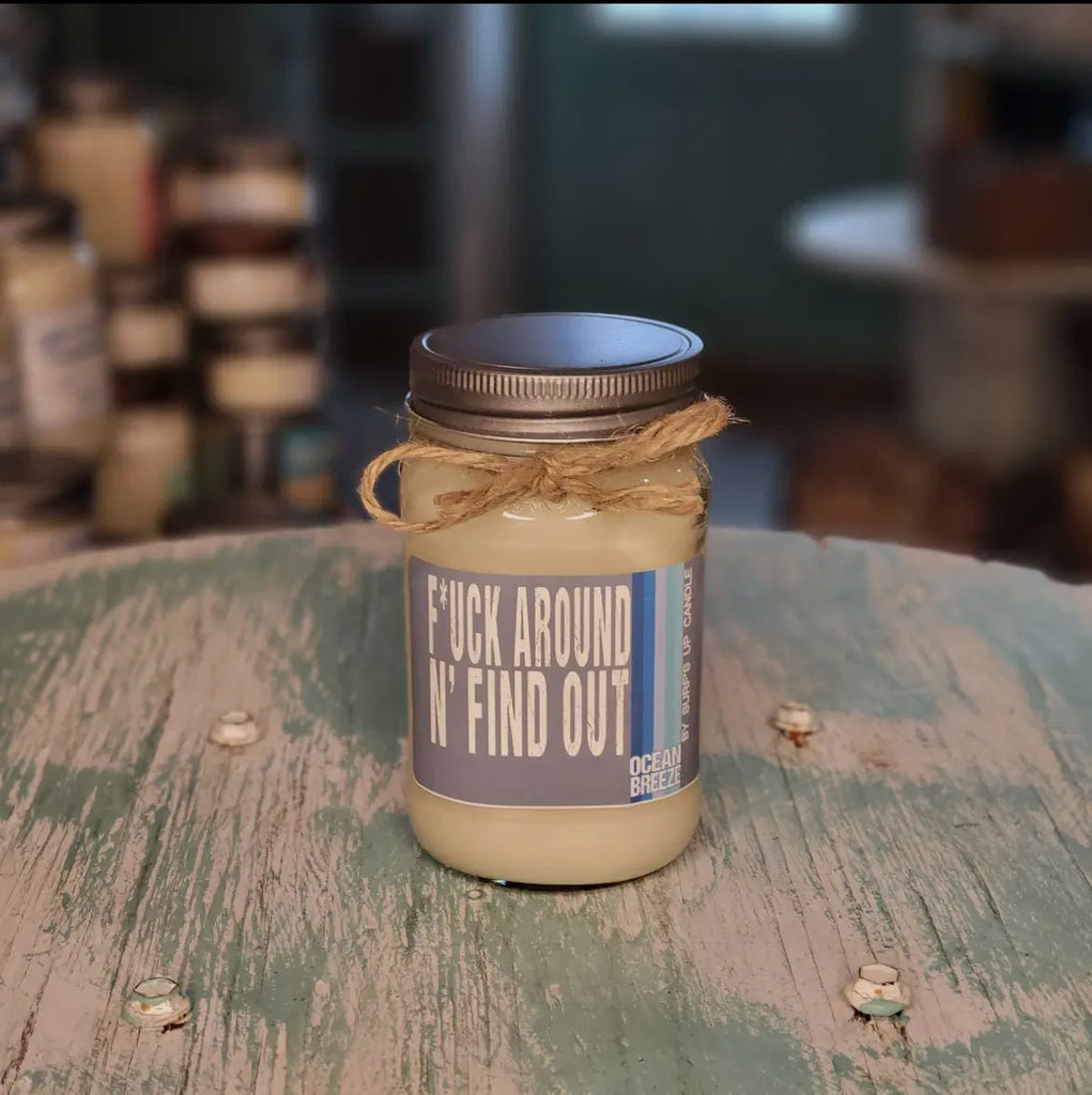 A Surfs Up Candles F*ck Around Ocean Breeze mason jar candle made from soy wax, complete with a silver lid and twine bow, rests on a rustic green table. The inviting scene is enhanced by a softly blurred background.