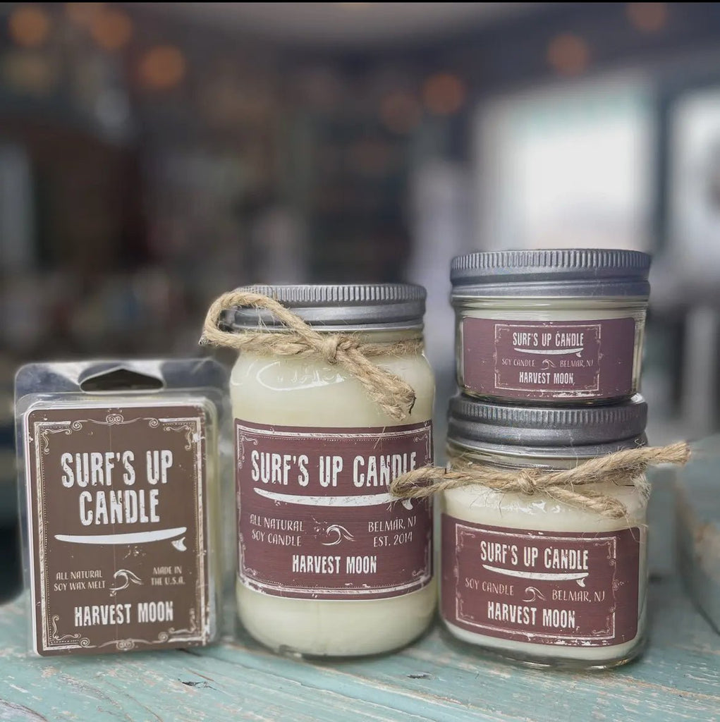 A display of Surfs Up Candles includes the Harvest Moon Mason Jar candle and a wax melt pack. These soy candles with metal lids and twine accents are set on a rustic wooden surface.