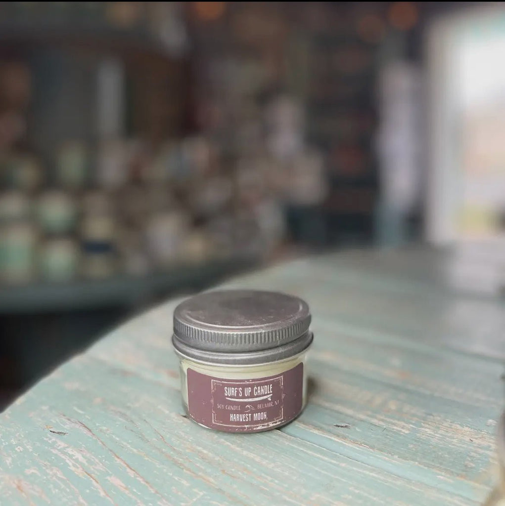 A Surf’s Up - Harvest Moon Mason Jar Candle from Surfs Up Candles, made of soy wax, sits on a weathered teal table. The background is blurred with various other candles.