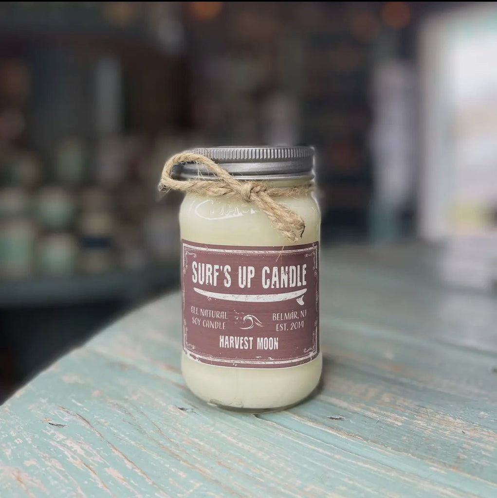 The Surf’s Up - Harvest Moon Mason Jar Candle by Surfs Up Candles features a warm Harvest Moon scent. Made with soy wax, it comes with a rustic brown label and a twine-wrapped lid, set on a weathered turquoise table against a blurred backdrop.